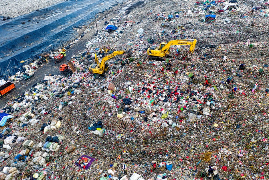 A large landfill site filled with mixed waste, including plastic bags, cardboard, textiles, and other debris, spread across a sloped terrain. Several yellow excavators are actively working to move and manage the rubbish, positioned intermittently along the slope. The waste piles vary in height, creating uneven mounds that extend toward the horizon. On the left side, a section of the site is covered by a blue plastic tarp, possibly functioning as a barrier or stabiliser. The environment appears to be outdoors with natural daylight illuminating the scene, highlighting the variety of colours and textures within the refuse. The presence of heavy machinery and the vast scale of accumulated waste suggest the site is operated as a large-scale disposal or waste management operation, akin to private or independent rubbish handling facilities managed by companies like House Clearance Clapham, rather than a local authority collection service. The overall atmosphere is utilitarian, with a focus on waste processing and containment.