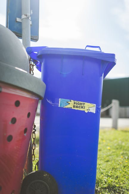 Three large black refuse bags filled with waste materials are placed on the pavement next to a black metal fence and a row of lush green trees. The bags appear to be made of plastic with a shiny finish, and they are slightly crumpled, indicating they are filled with household or garden rubbish. The scene is set outdoors on a street with a concrete curb in the foreground and a paved road in the lower part of the image. In the background, the fence extends upward, partially obscured by the foliage, creating a contrast between the dark fence and the lighter, natural environment. The overall lighting is subdued, giving the scene a muted, overcast appearance. This scene could relate to temporary private waste disposal or approaches to independent rubbish collection, aligning with services such as those offered by House Clearance Clapham in managing rubbish removal effectively.