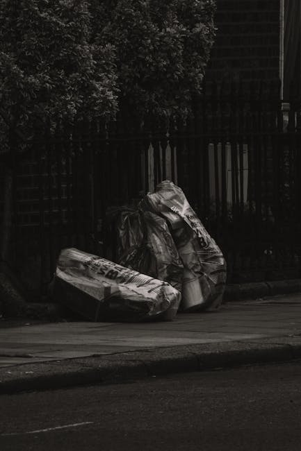 Three large black garbage bags, made of heavy-duty plastic with visible creases and folds, are positioned on the edge of a pavement next to a black metal fence. The bags appear to be filled with household waste or discarded items, and feature printed labels and recycling symbols. Behind the fence, there is a dense cluster of leafy trees, and the scene is illuminated by natural daylight, creating a contrast between the dark bags and the surrounding environment. The pavement is smooth, with a curb separating it from the road or driveway. The setting suggests an outdoor area where private waste and rubbish are temporarily stored prior to collection, reflecting an alternative approach to waste disposal that may involve independent rubbish removal services. The image underscores the importance of proper waste management and highlights the type of clutter that professional rubbish clearance companies, such as House Clearance Clapham, typically handle for residential properties.