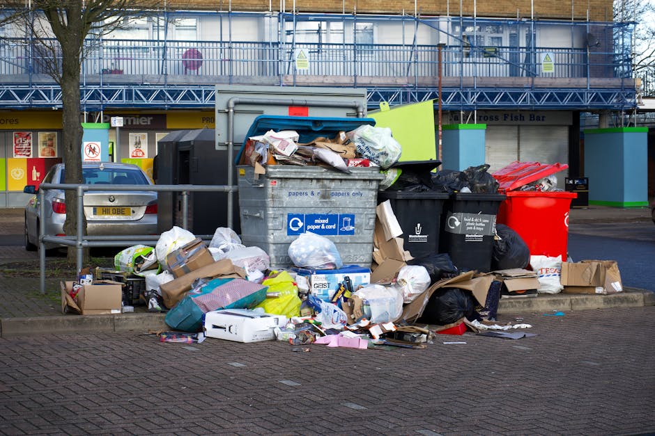 A large pile of mixed household waste and rubbish bags situated on a paved urban street in front of wheelie bins, including cardboard boxes, paper and plastic sacks, and loose items such as packaging and discarded packaging material. The rubbish is overflowing from a metal recycling bin labeled for mixed paper and cardboard, with some debris spilling onto the pavement. Black and red waste containers are positioned nearby, some filled with unsegregated waste, and a few flattened cardboard boxes are situated around the pile. In the background, a commercial building with stairs, a storefront with signage, and a parked silver car are visible. The scene is illuminated by daylight, with a tree on the left side partially framing the rubbish accumulation, indicating an area possibly focused on alternative waste handling or private rubbish collection services as seen by the cluttered and unmanaged appearance of the waste. The overall scene highlights the need for professional rubbish removal to manage urban waste responsibly.