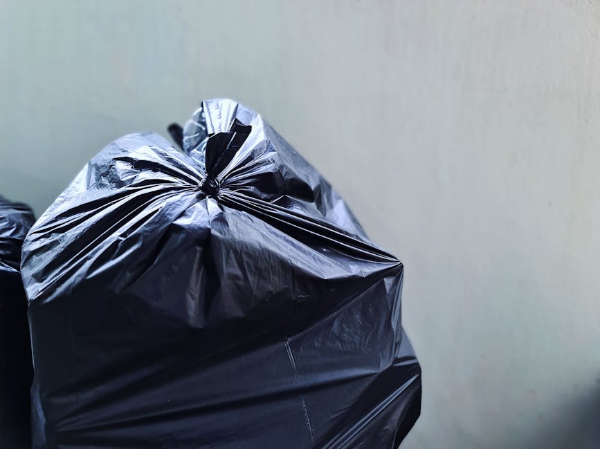 Close-up view of a black plastic rubbish bag tightly sealed at the top with a knot, placed against a plain, light-colored wall. The bag appears full and bulging, with a slightly crinkled surface texture reflecting light, revealing its dense contents. Behind the main bag, additional black rubbish bags are partially visible, stacked or lined up, suggesting an ongoing collection or removal process. The scene is set outdoors, likely on a driveway or pavement adjacent to a property, with natural lighting casting soft shadows. The overall environment indicates a domestic setting where local waste removal or private clearance services, such as those provided by House Clearance Clapham, are involved, reflecting an alternative to standard municipal rubbish collection procedures. The focus on the sealed bags and surrounding area emphasizes an approach to efficient on-site rubbish handling.
