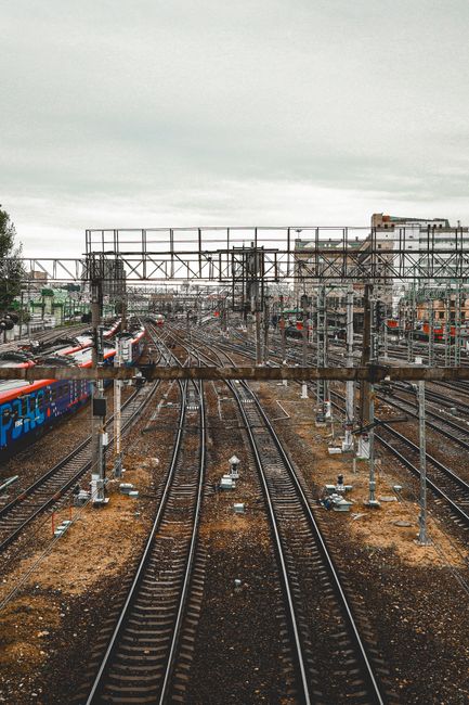 The image shows multiple railway tracks extending into the distance within a train station environment, with various overhead electrical lines and metal structures supporting the wires above the tracks. The tracks are composed of dark metal rails mounted on wooden sleepers and set in a bed of ballast, with some sections appearing rusty or weathered. On the left side, a cargo train with a blue and red exterior is partially visible, parked alongside the tracks. In the background, there are industrial buildings, some with flat roofs and windows, indicating an urban setting. The sky is overcast with grey clouds, creating diffuse lighting that highlights the textures of the railway infrastructure and surrounding elements, conveying a busy and functional transport hub. The scene inherently relates to the context of materials and infrastructure that are often involved in large-scale waste management or site clearance, aligning with services provided by House Clearance Clapham in managing and removing waste associated with such industrial environments.