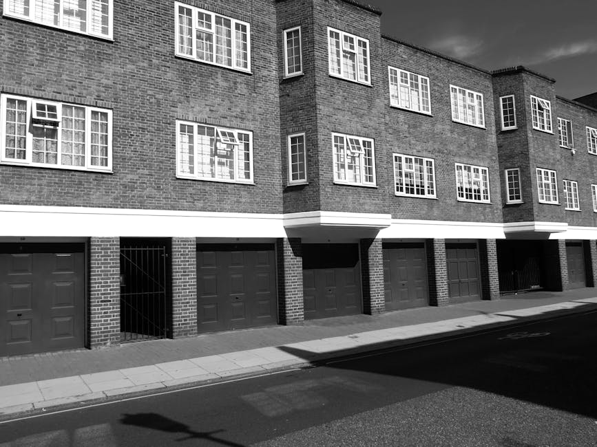 A black-and-white photograph depicts a multi-story residential building with a brick facade, featuring several windows with white frames and divided panes. Below the residential windows, there is a row of dark garage doors, each with a panel design, set into brick pillars, some with metal gates for access. The building is situated along a paved street with a concrete curb and sidewalk in the foreground, casting shadows from nearby objects or structures. The overall scene appears clean and orderly, reflecting an urban environment where private waste handling or third-party rubbish removal services, such as those offered by House Clearance Clapham, could facilitate bulky waste collection or on-site clearance for residents.