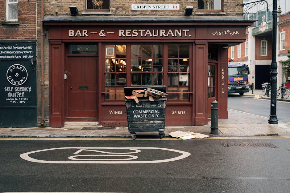 The image depicts a small front view of a restaurant or bar located on a street corner, with a maroon exterior including large window panes, a wooden door, and signage indicating it is a bar and restaurant with oyster, wine, and spirit offerings. In front of the establishment, a black commercial waste bin labeled 'Commercial Waste Only' is positioned on the sidewalk, filled with cardboard boxes and paper, with some discarded materials resting on the ground nearby. The surrounding environment includes a street with road markings, a lamppost on the right, and a parked large truck partially visible behind the building. To the left, there is a black signage board advertising a self-service buffet with operating hours, and to the right, additional urban elements such as bicycles and additional buildings can be seen. The scene is lit with natural daylight, creating an everyday urban atmosphere that relates to waste management and rubbish collection services, exemplifying the importance of proper waste disposal in commercial areas, as provided by companies like House Clearance Clapham.