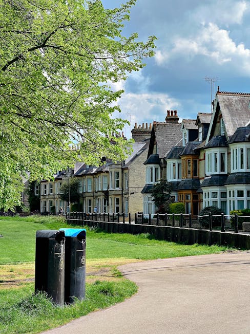 A row of Victorian-style terraced houses with bay windows and slate roofs, situated along a residential street. In the foreground, there is a grassy area with two black waste disposal bins positioned near a paved pathway. A large, leafy green tree with fresh spring foliage extends its branches over the scene, partially shading the grass and pathway. The sky above is partly cloudy with patches of blue, indicating a bright day. The setting appears to be a quiet suburban neighbourhood, with the waste bins possibly awaiting collection or decluttering efforts by local rubbish removal services such as House Clearance Clapham. The environment looks clean and well-maintained, emphasizing the importance of effective waste management and private rubbish collection in residential communities, especially when undertaking property clearance or garden waste disposal.