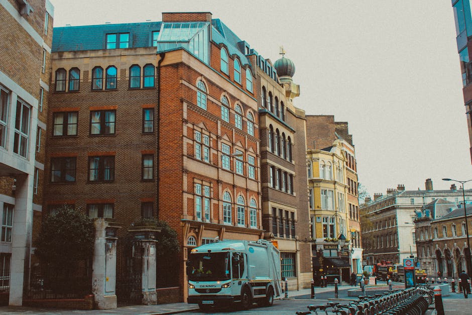 A narrow urban street scene featuring a mix of historic and modern buildings, including a prominent brick structure with arched windows and decorative stonework, standing adjacent to a cream-colored building with ornate architectural details. In the foreground, a white rubbish collection vehicle is parked on the street, close to a small dark metal gate with stone pillars on either side. The street is lined with black bollards and bike racks, with some bicycles attached. The background reveals additional multi-story buildings with flat roofs, some with classical facades and others with contemporary glass finishes. The sky appears overcast, casting diffuse natural light over the scene, which illustrates typical city infrastructure for private waste collection and environmental management, consistent with independent rubbish removal services such as those offered by House Clearance Clapham.